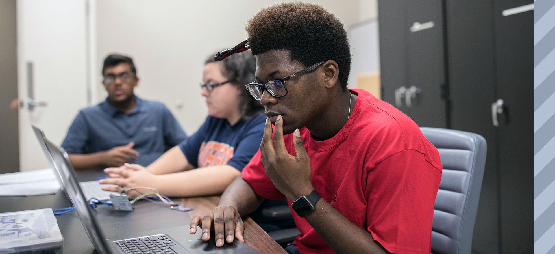 Young African-American man in foreground looks intently into laptop screen. In the background are a female and male student.