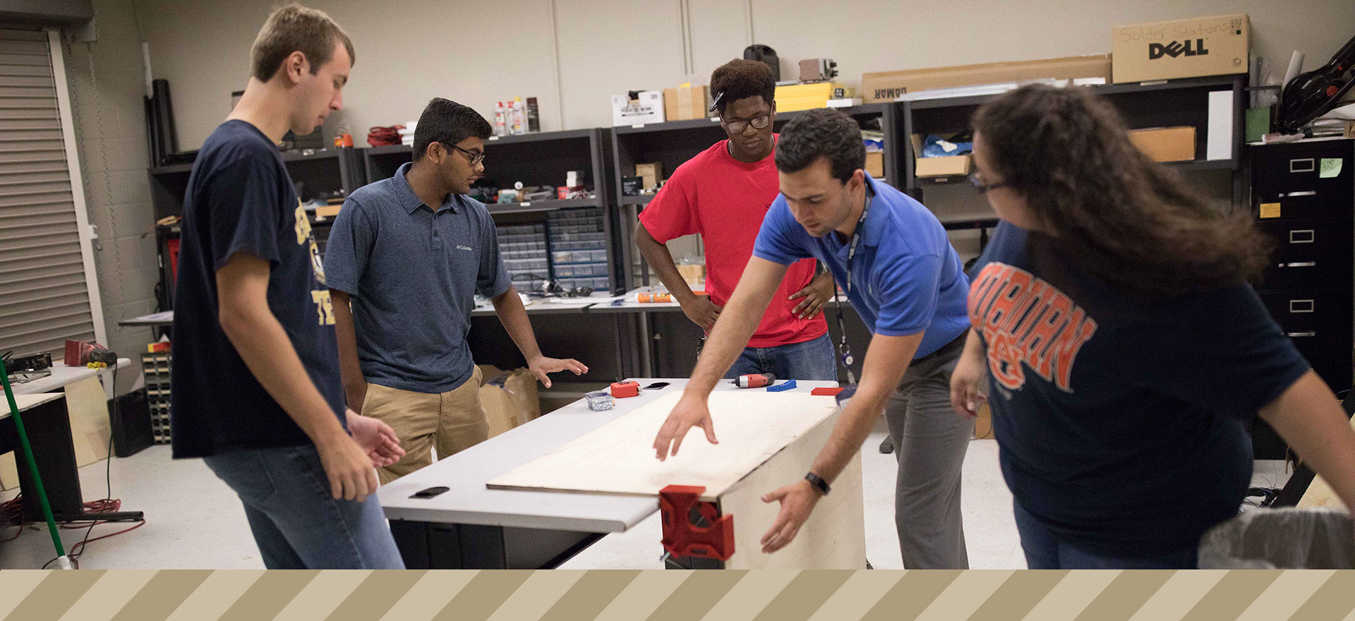 Four students gathered around their mentor, who is demonstrating a wood clamp on a box in the lab.