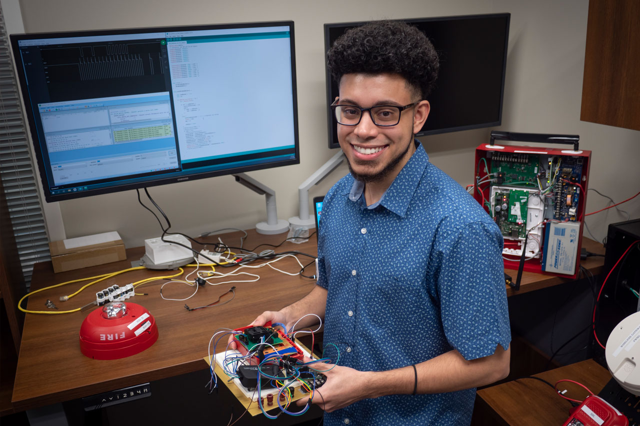 Young male student stands in front of computer screen while holding parts of electronics.