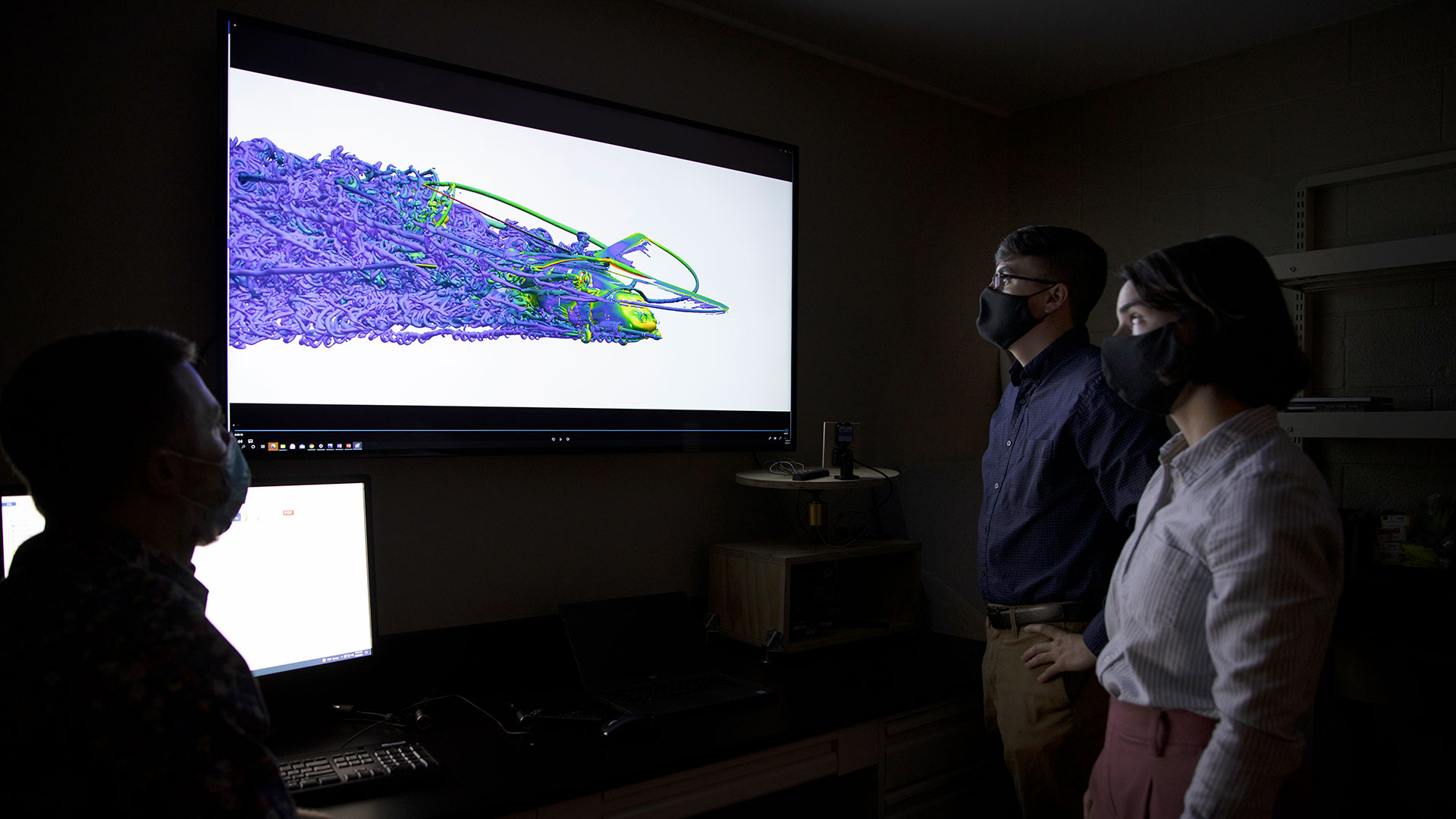 Two male and one female researcher in dark room with large video display showing brightly colored computer simulation of air flow around helicopter 