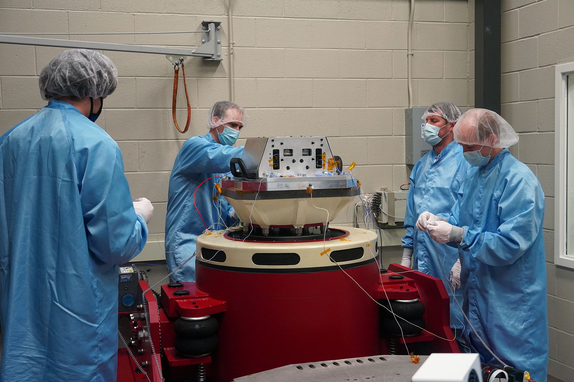 Three researchers dressed in gowns, masks, and hairnets assemble device in lab.