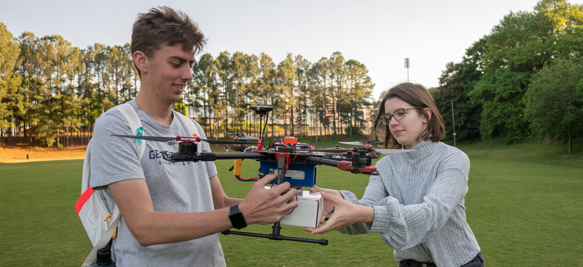 Male student holds drone while female student adjust package the drone carries. The scene is a green field outdoors, in the afternoon, with slightly overcast sky.