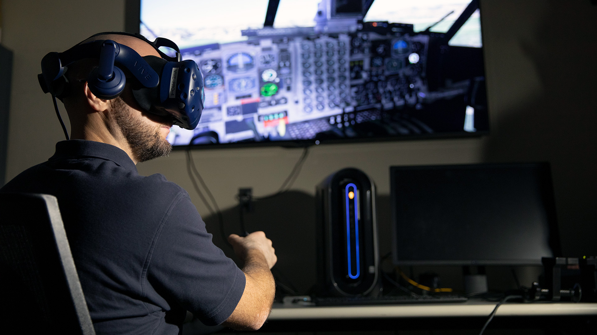Male researcher sitting at table containing FITRE hardware, there is a large video screen, and he is wearing VR goggles and holding a control stick
