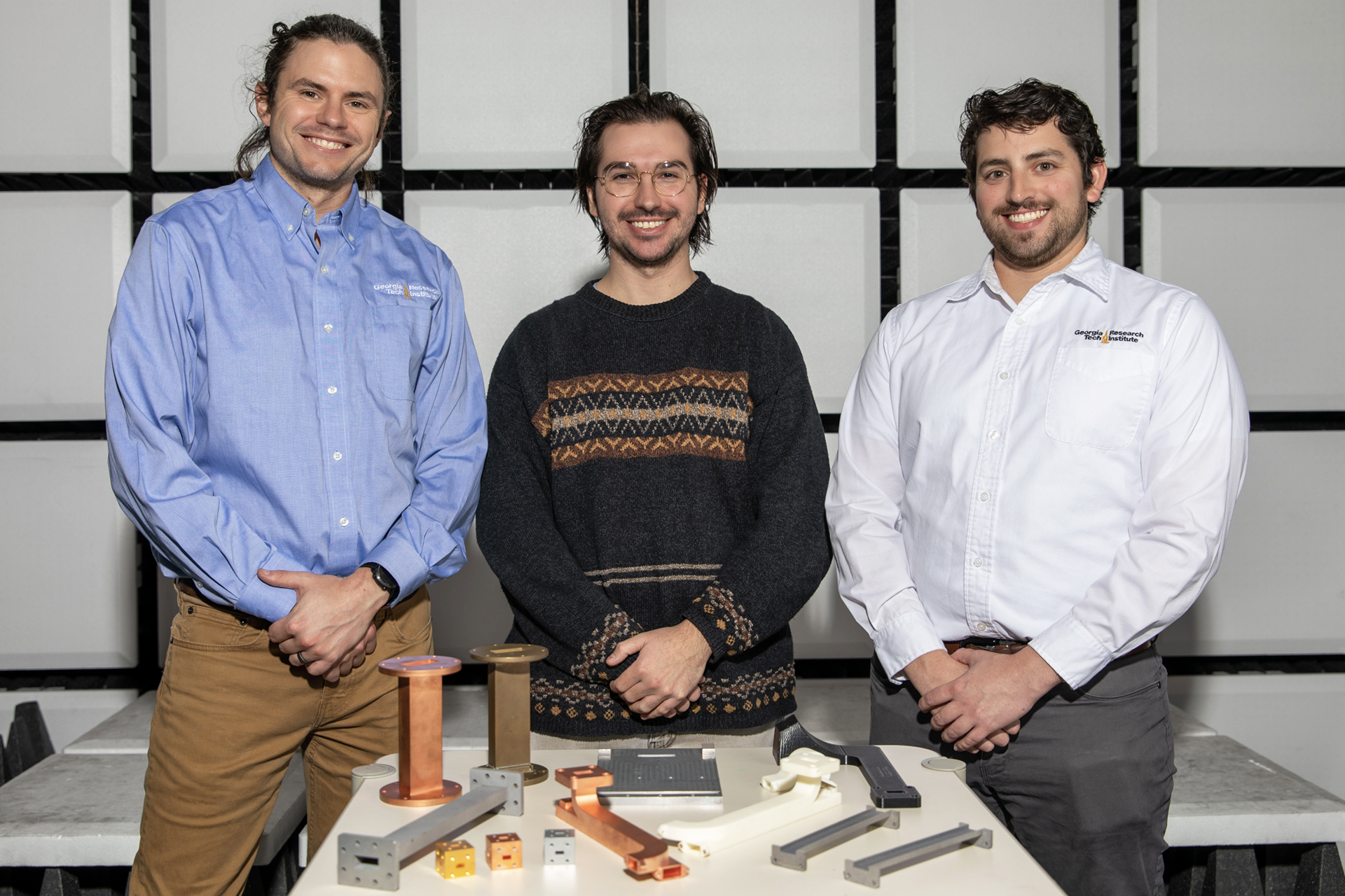 Three male researchers pose behind small table containing various 3D printed components