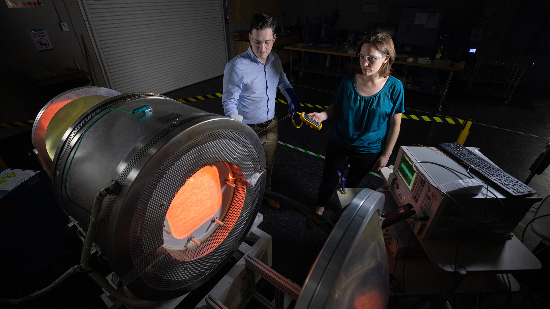 Two research standing behind large furnace device glowing red.
