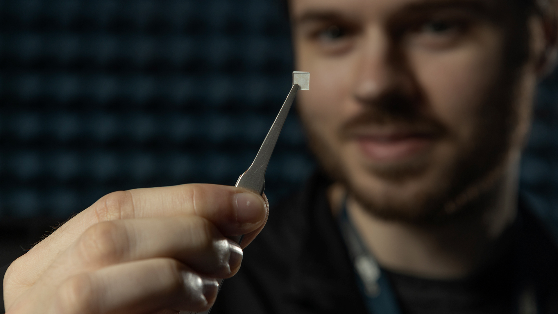Researcher holds tweezers with small chip up to camera. Room is dark and researcher's face is somewhat obscured.