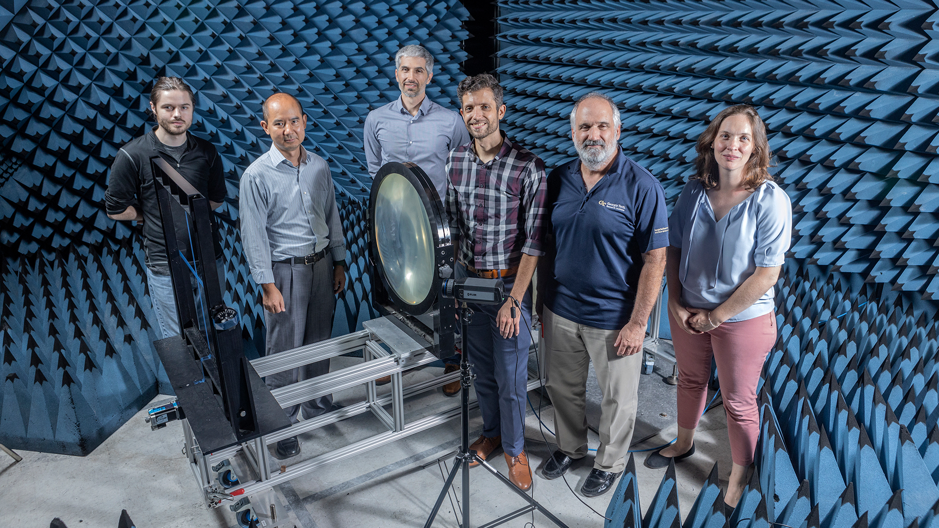 Five male and one female researcher stand in a group behind the testing device inside the dark blue anechoic chamber.