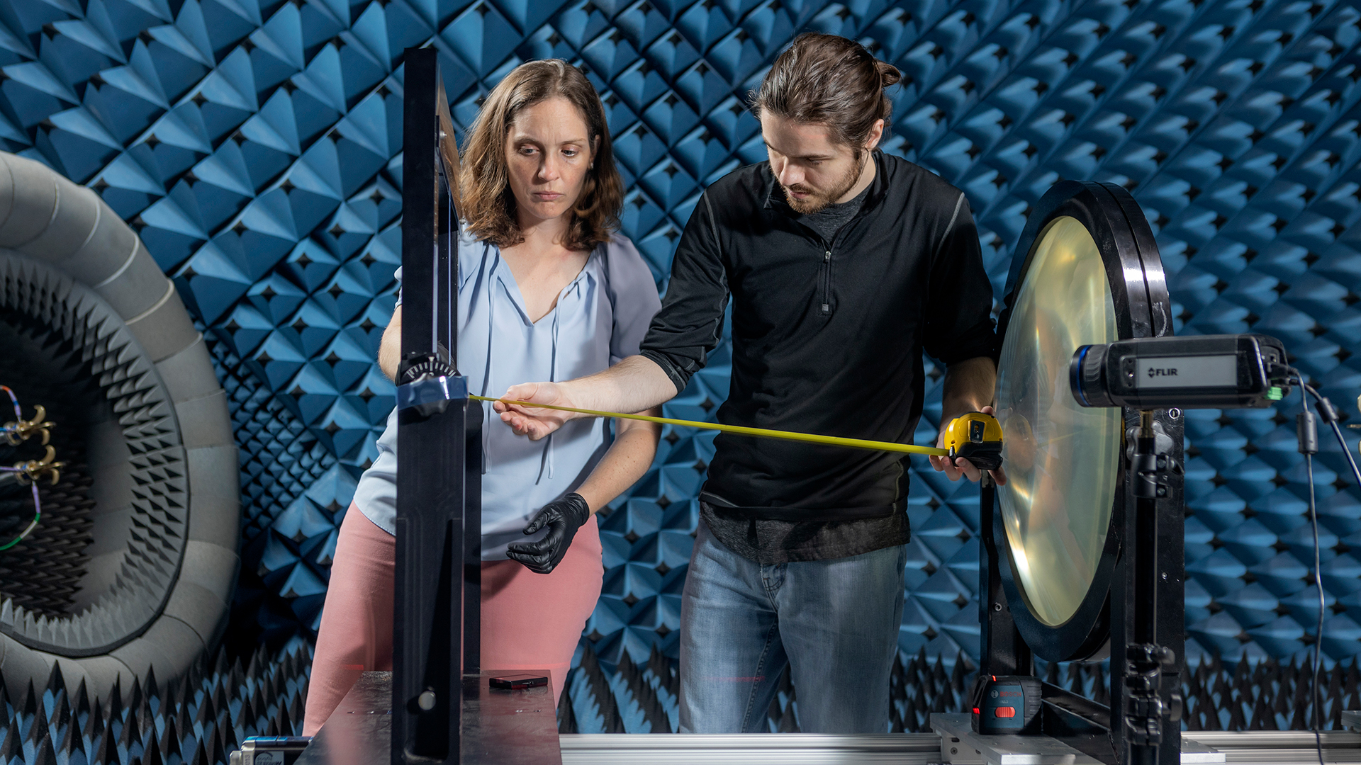One male and one female researcher in the dark blue anechoic chamber measuring distance between two parts of the testing device.