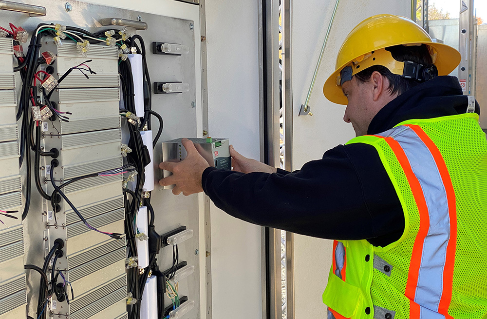 Worker in hardhat and reflective vest standing in front of a panel of wiring, placing a small box inside the device.