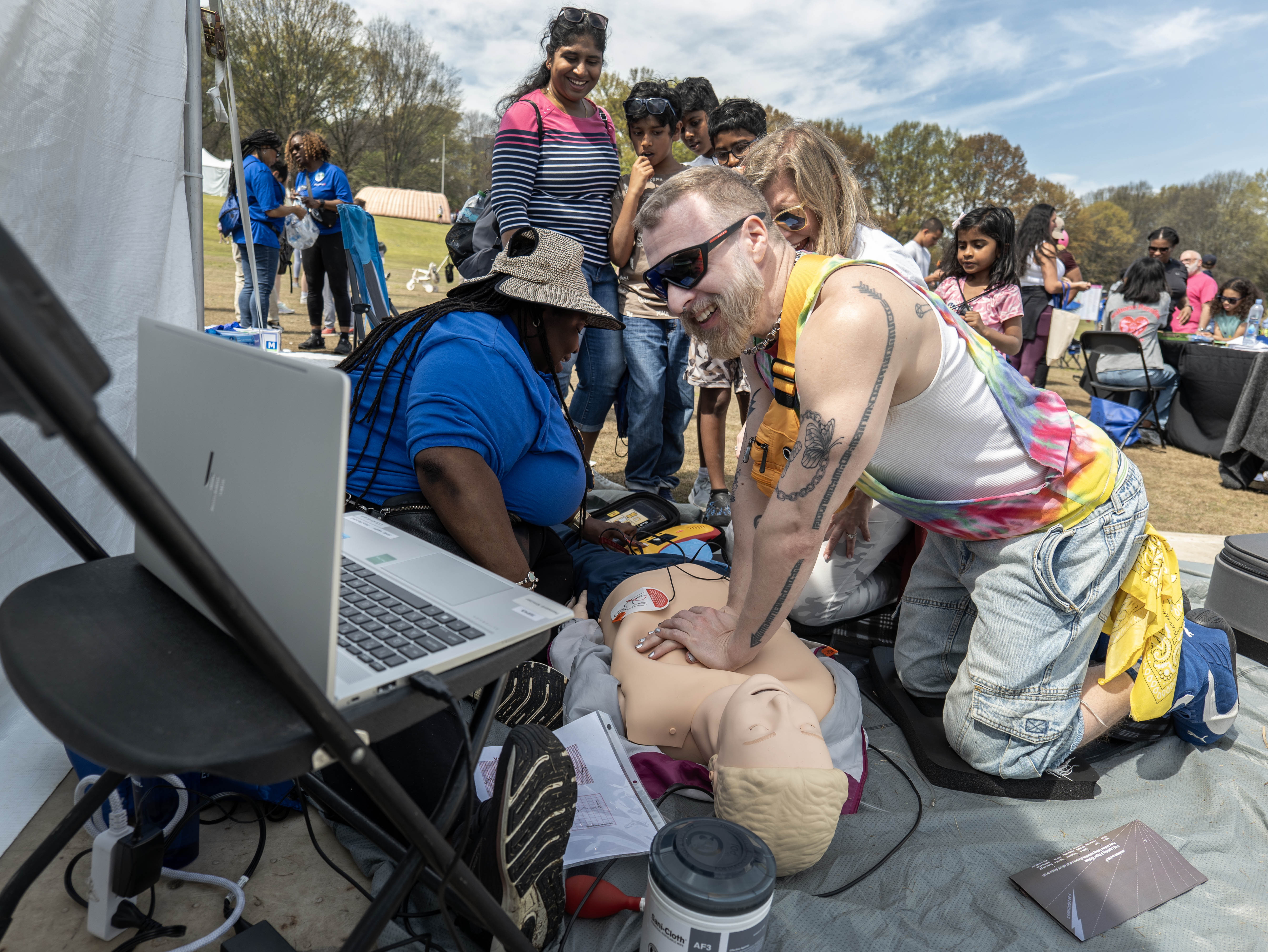 A man performing CPR chest compressions on a dummy with a woman in front supevising. Groups of people including women and children in the background watching.