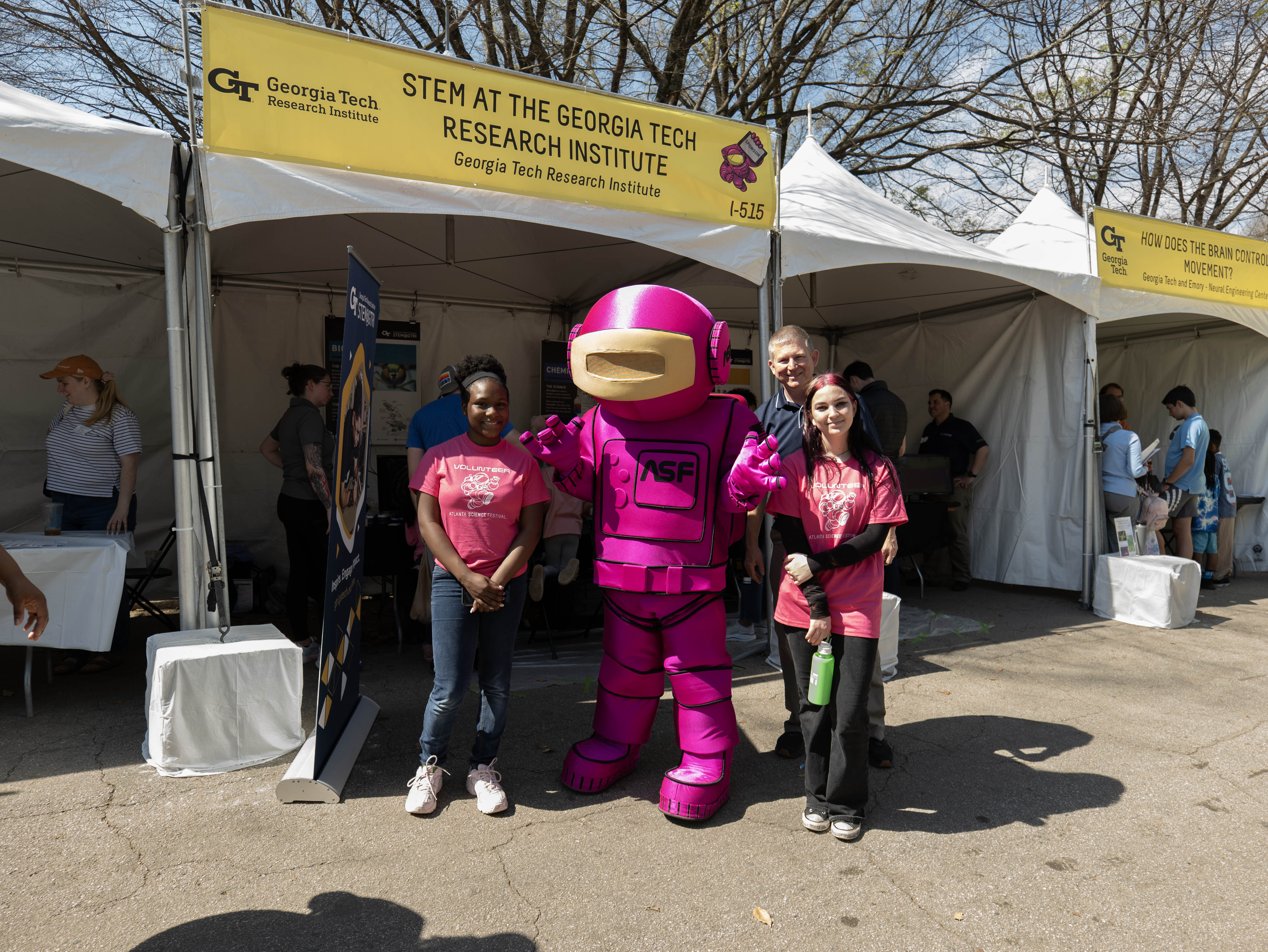 Three people pose with a person in a bright pink astronaut mascot costume in front of a Georgia Tech Research Institute STEM tent at an outdoor event.