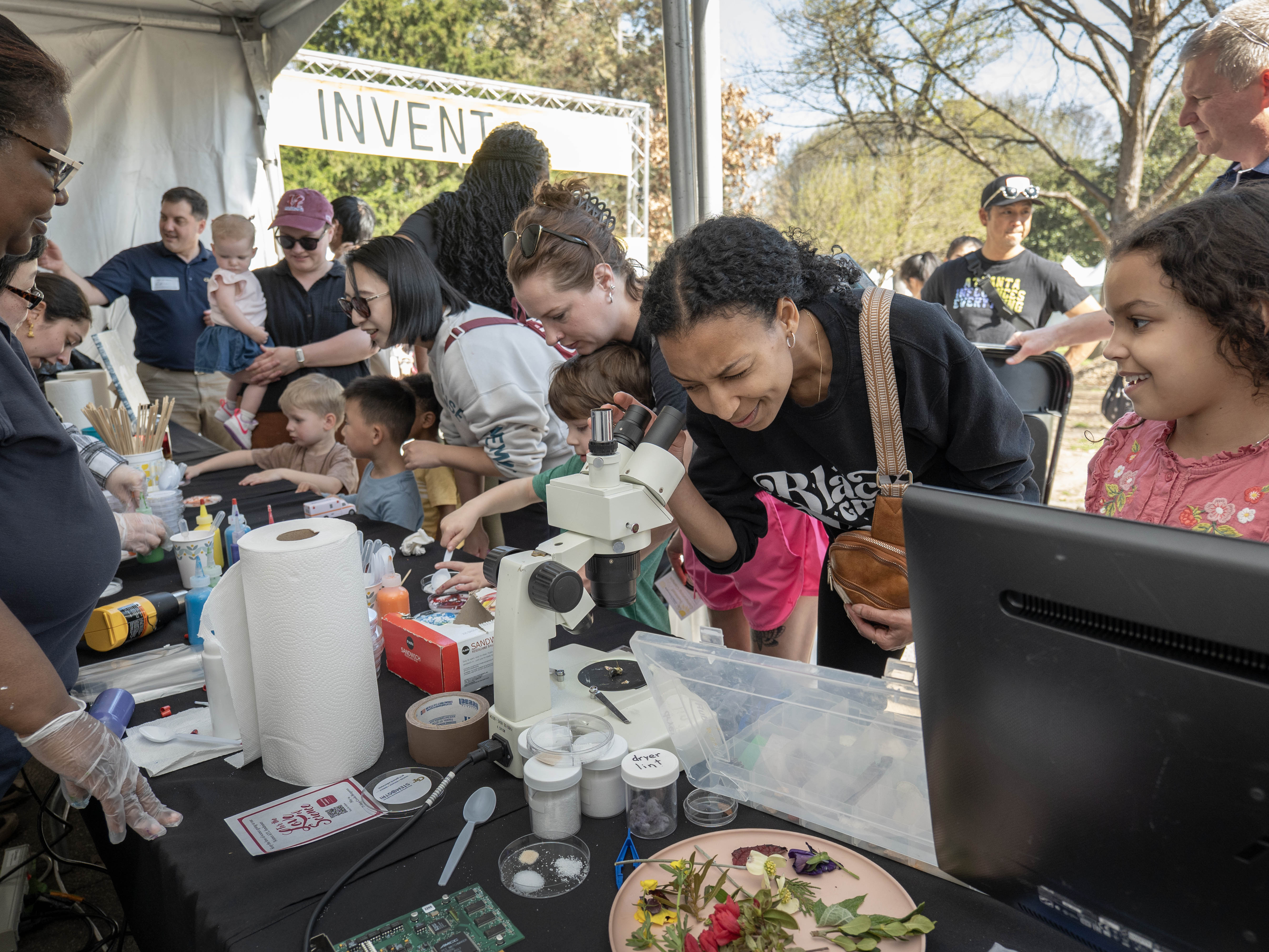 Children and adults explore science activities with microscopes and specimens at an outdoor STEM event under a tent with an 'INVENT' sign in the background..