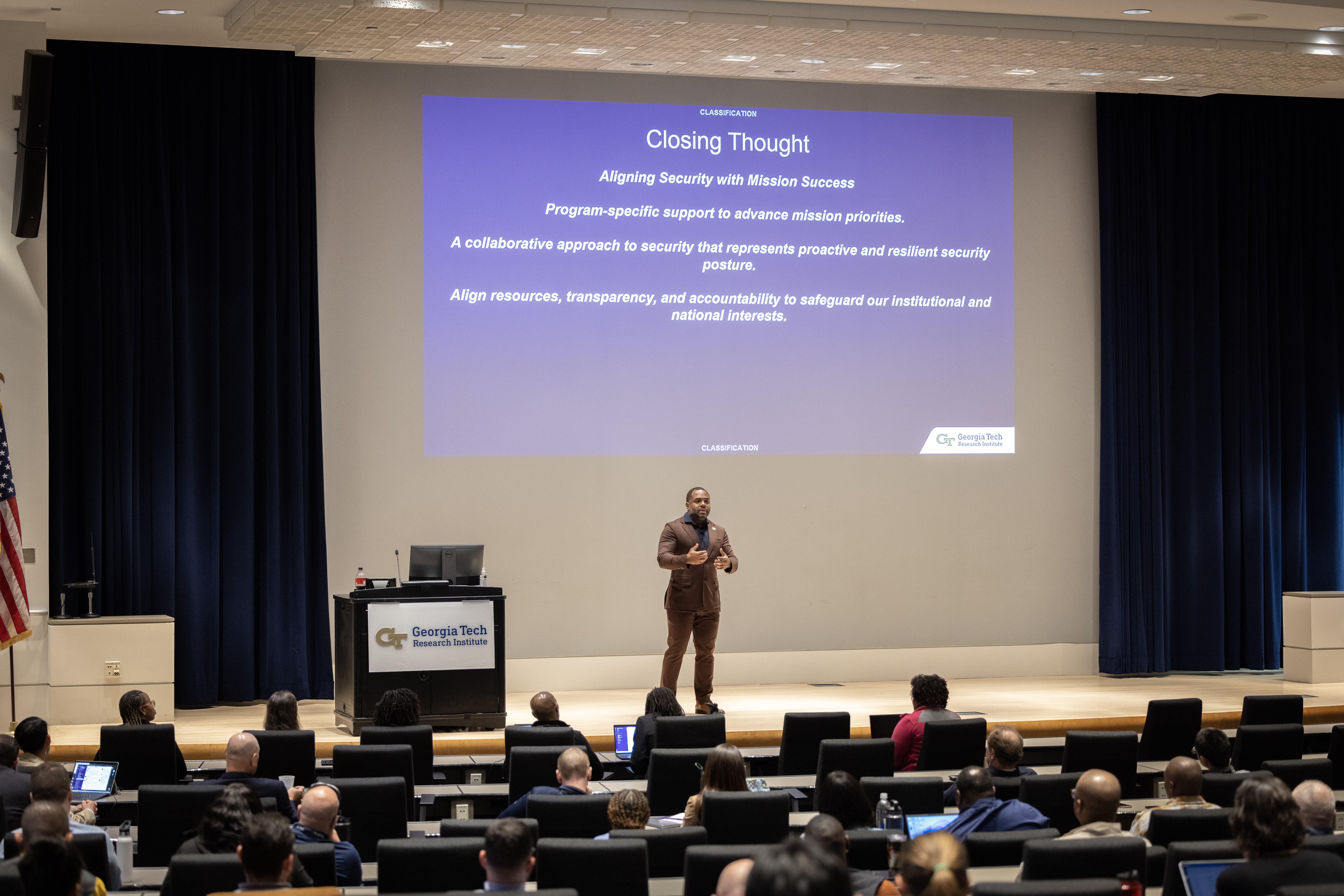 Jonathan Brown presents on stage at GTRI town hall with 'Closing Thought' slide about aligning security with mission success displayed behind him.