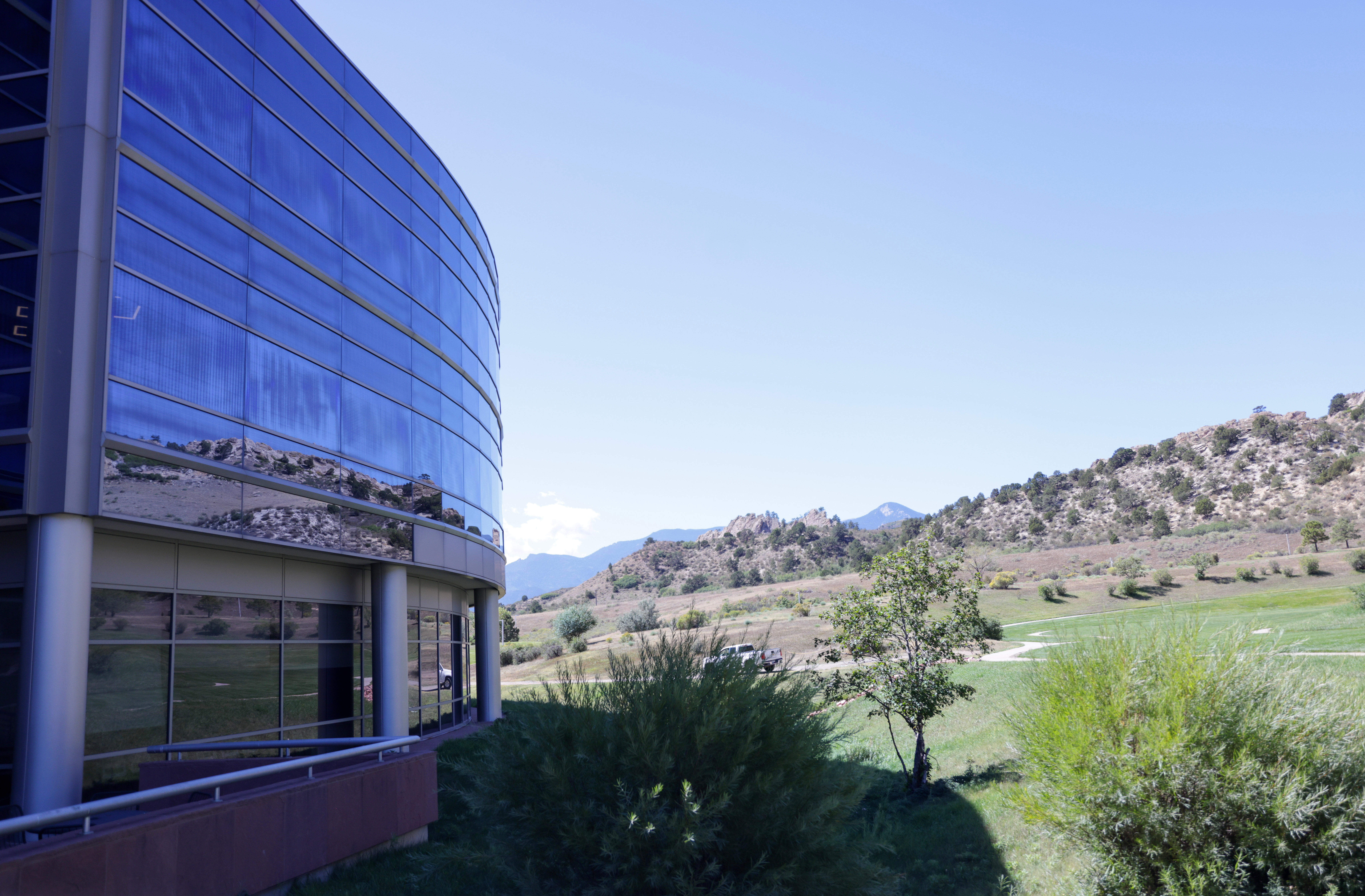 Modern curved glass office building with blue-tinted windows overlooking a green valley with mountains in the distance under clear sky.