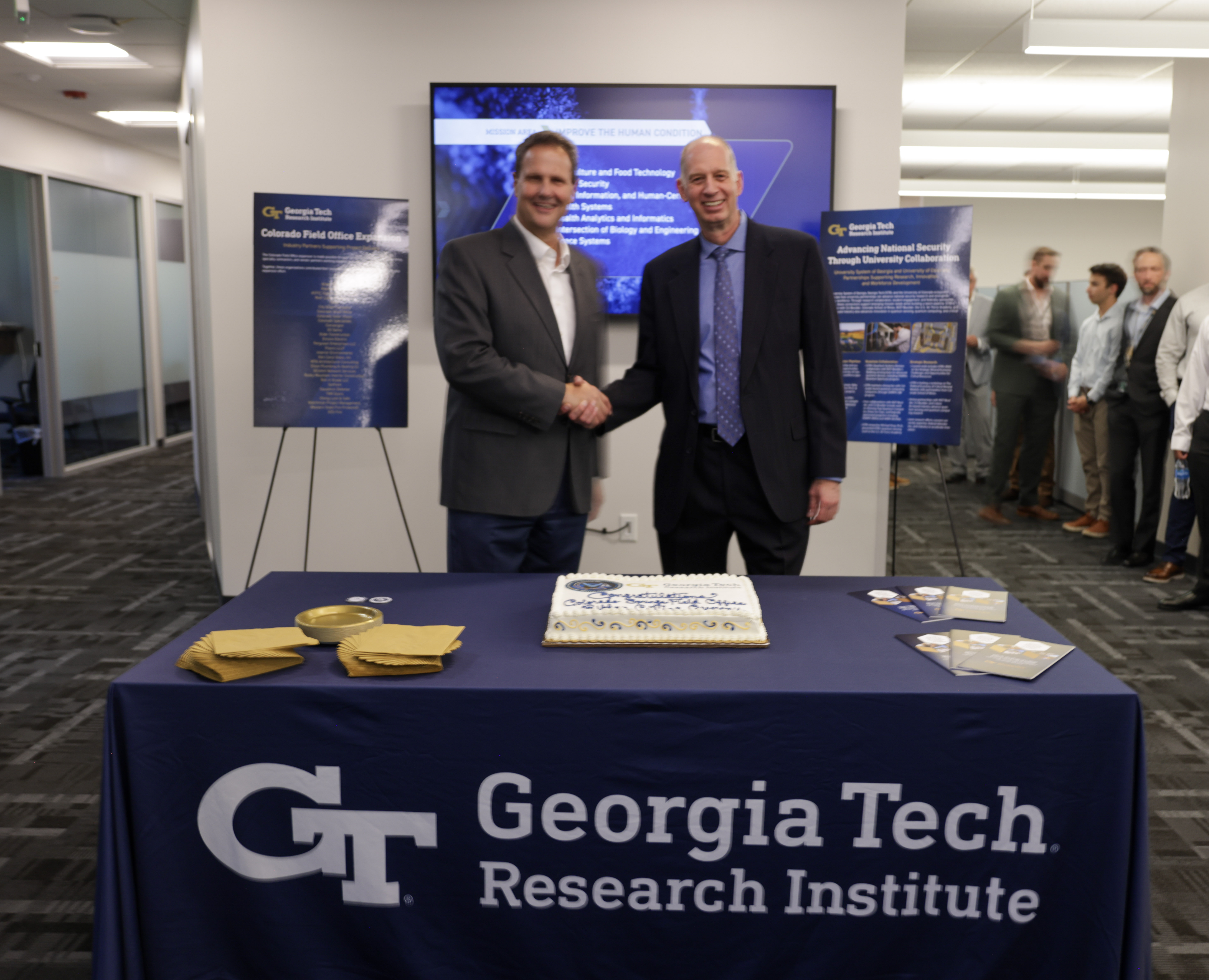 Two men in business suits shake hands behind a Georgia Tech Research Institute table with celebration cake, at an office opening ceremony event.
