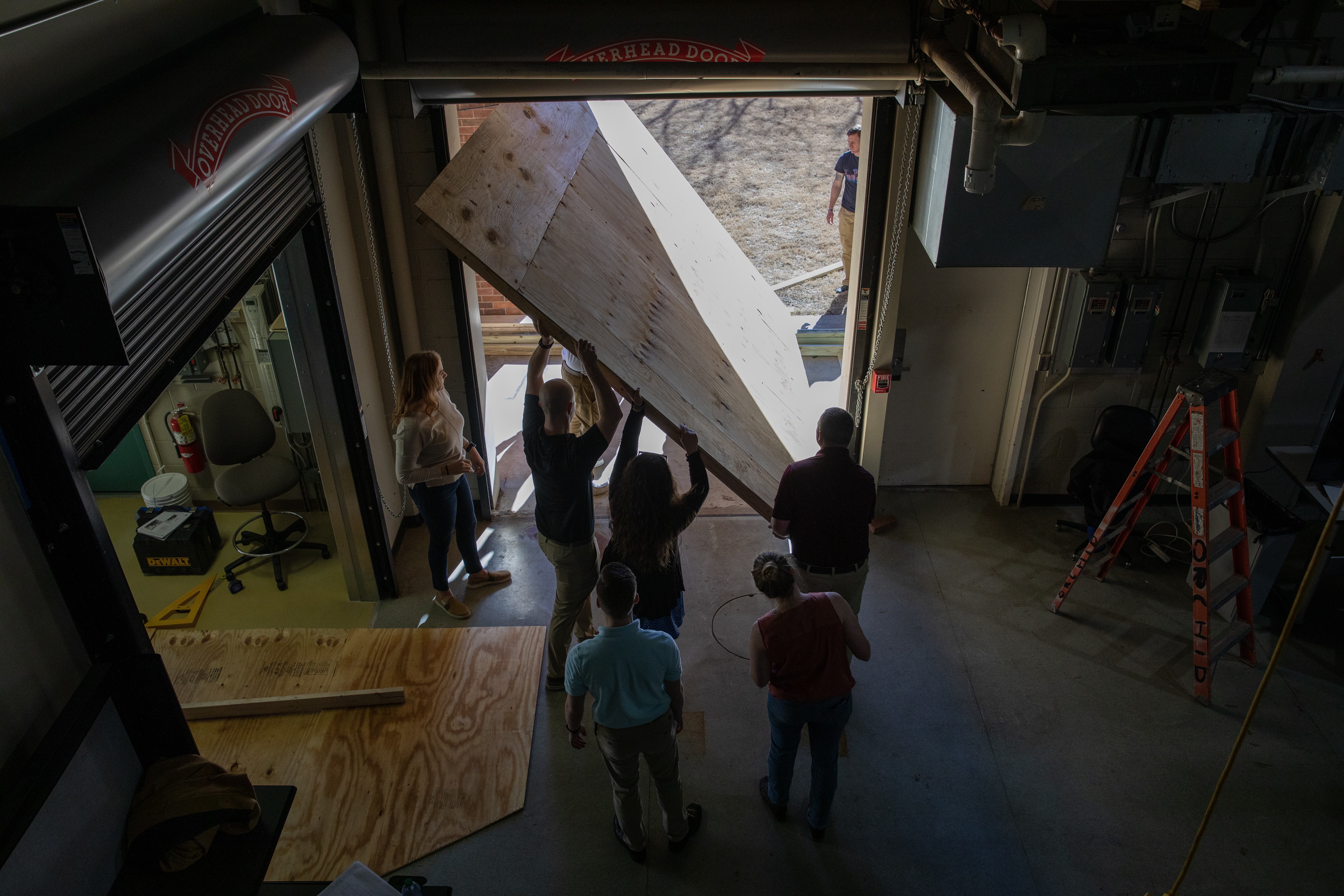A group carrying a large piece of wood outside of the workshop