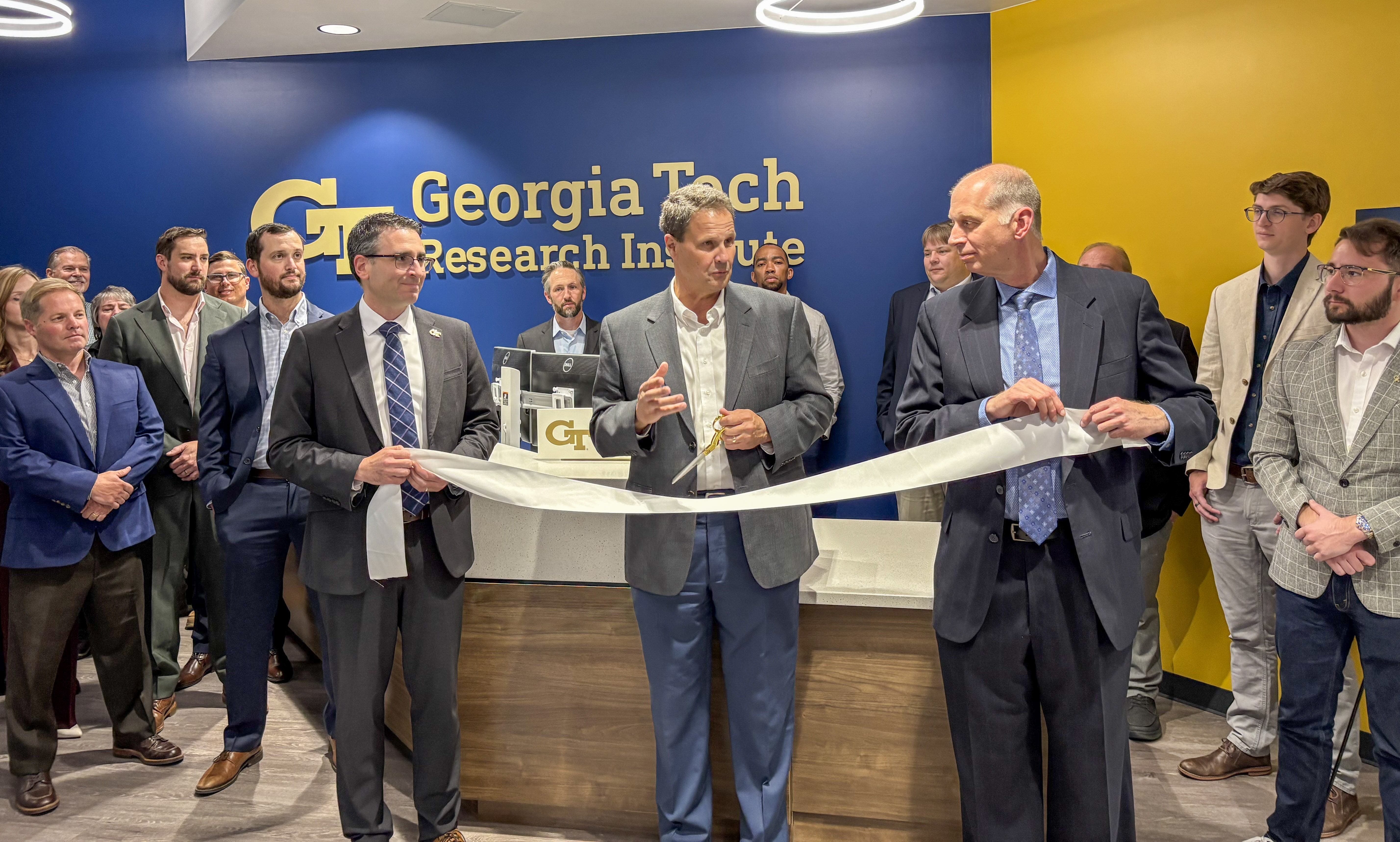 Three men in suits prepare to cut ribbon at Georgia Tech Research Institute office opening ceremony, surrounded by attendees.