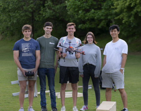 Five students standing in a row in a field smiling and looking at camera. Student in center is holding a drone.