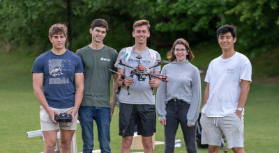 Five college students (4 males, 1 female) standing together outdoors, in a row.