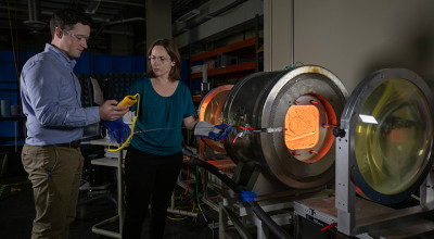 Two researchers stand near a large metal instrument with center glowing red hot