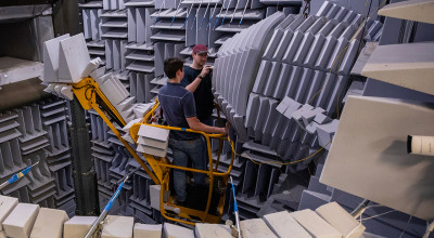 Two male researchers standing in cherry picker lift,  in large acoustic testing room.