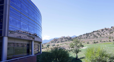 Modern curved glass office building with blue-tinted windows overlooking a green valley with mountains in the distance under clear sky.