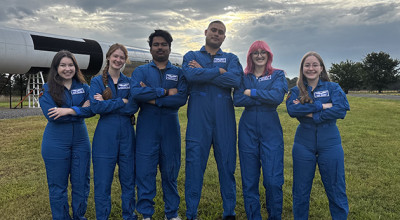 Six diverse team members in blue flight suits stand with arms crossed on grass field under dramatic cloudy sky, with aircraft visible behind them.